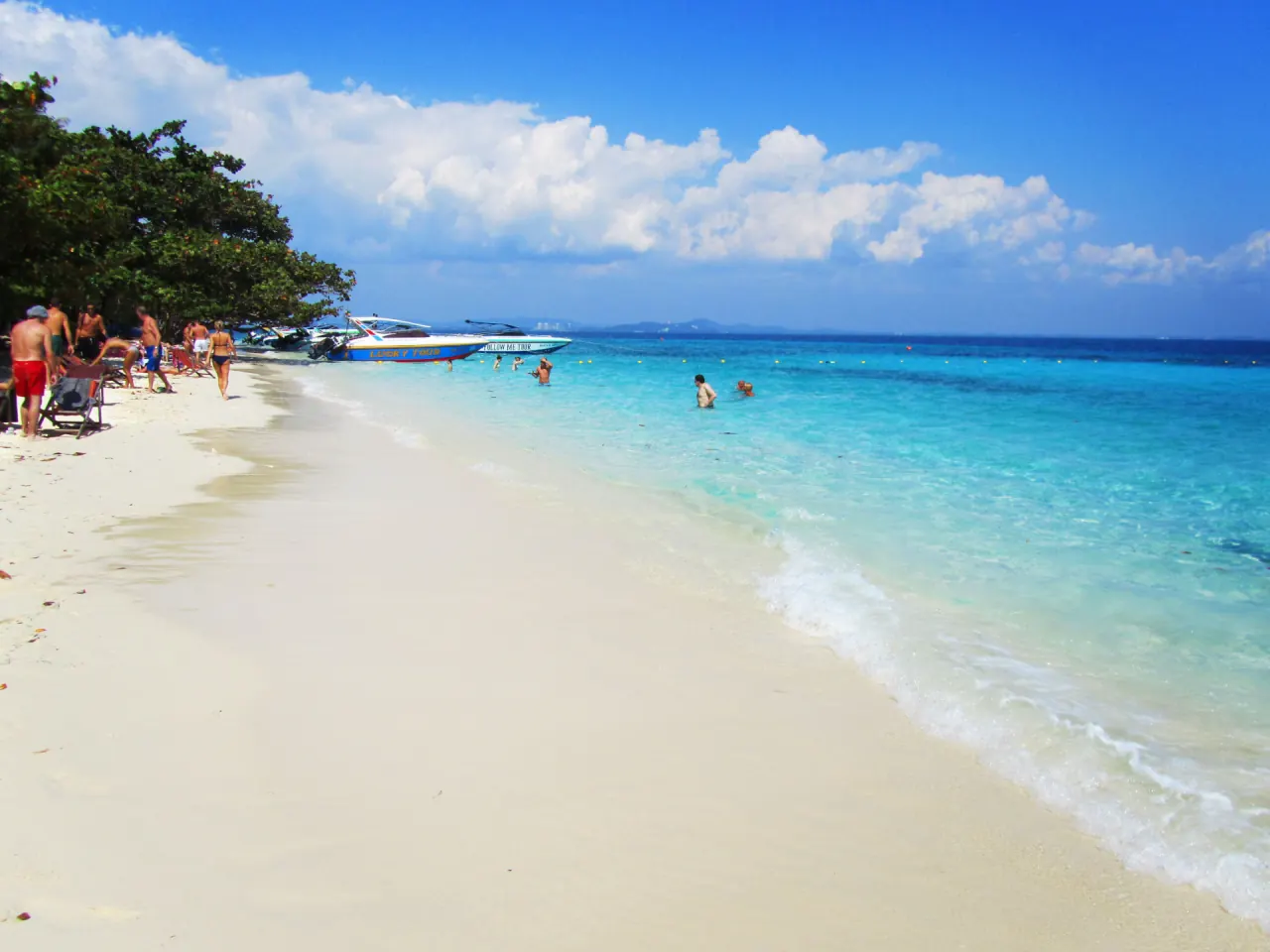 Strand med hvit sand og turkist vann under blå himmel, med fargerike speedbåter ved vannkanten og folk som bader og går langs stranden